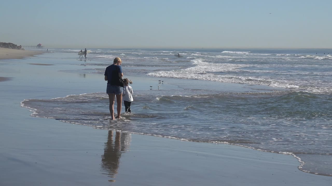 Adult and child stand together in shallow water at Huntington Beach, California, while a wider sheet of surf moves in from the right side of the frame.