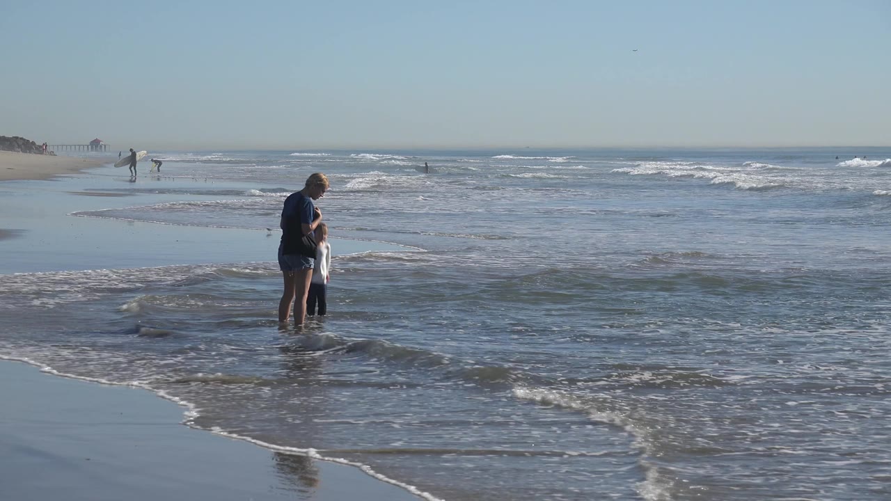 A woman and child stand farther right in ankle-deep water at Huntington Beach, with layered white surf behind them and a clear sky above the horizon.