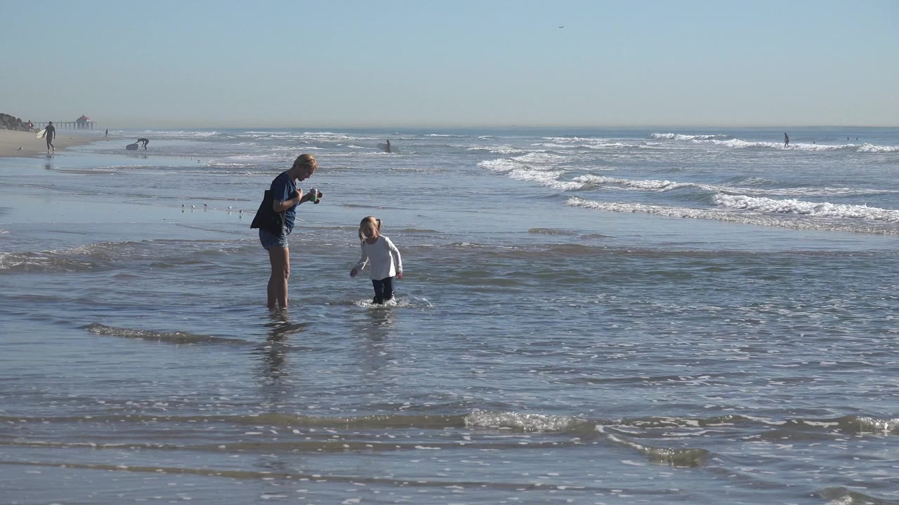 At Huntington Beach, California, an adult woman stands near a child in a white shirt as a small wave reaches their legs on wet sand.