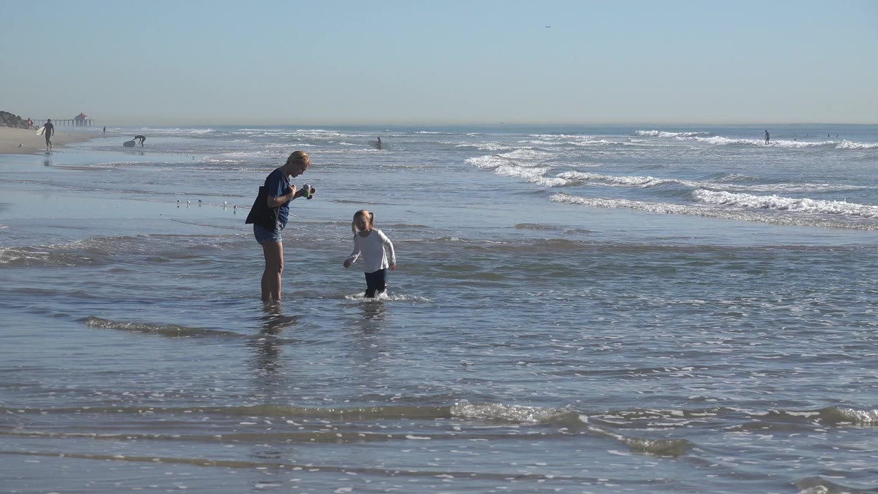 At Huntington Beach, California, the child in a white shirt faces the adult woman while white surf rolls in behind them under a clear sky.