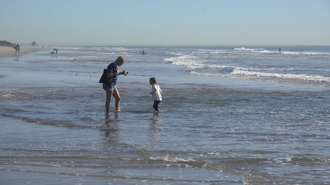 At Huntington Beach, California, the adult woman leans toward the child in shallow water while low waves break across the midground.