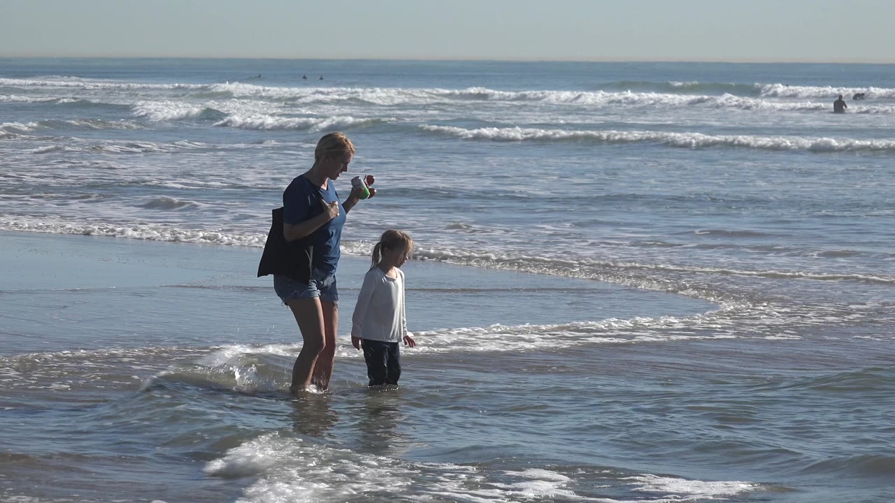 Adult woman holding a small item while a child stands beside her in surf.