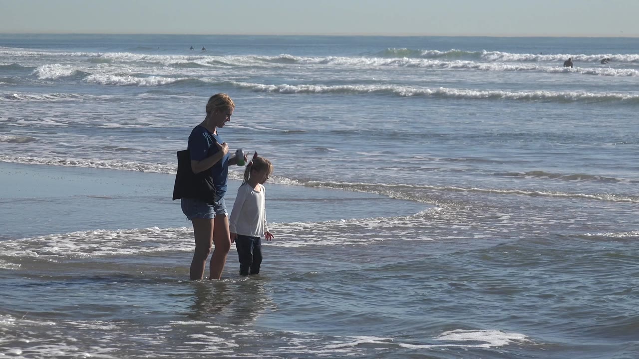 The adult woman and child stand closer together in Huntington Beach surf, with the child turned toward the adult and foam spreading over the wet sand.