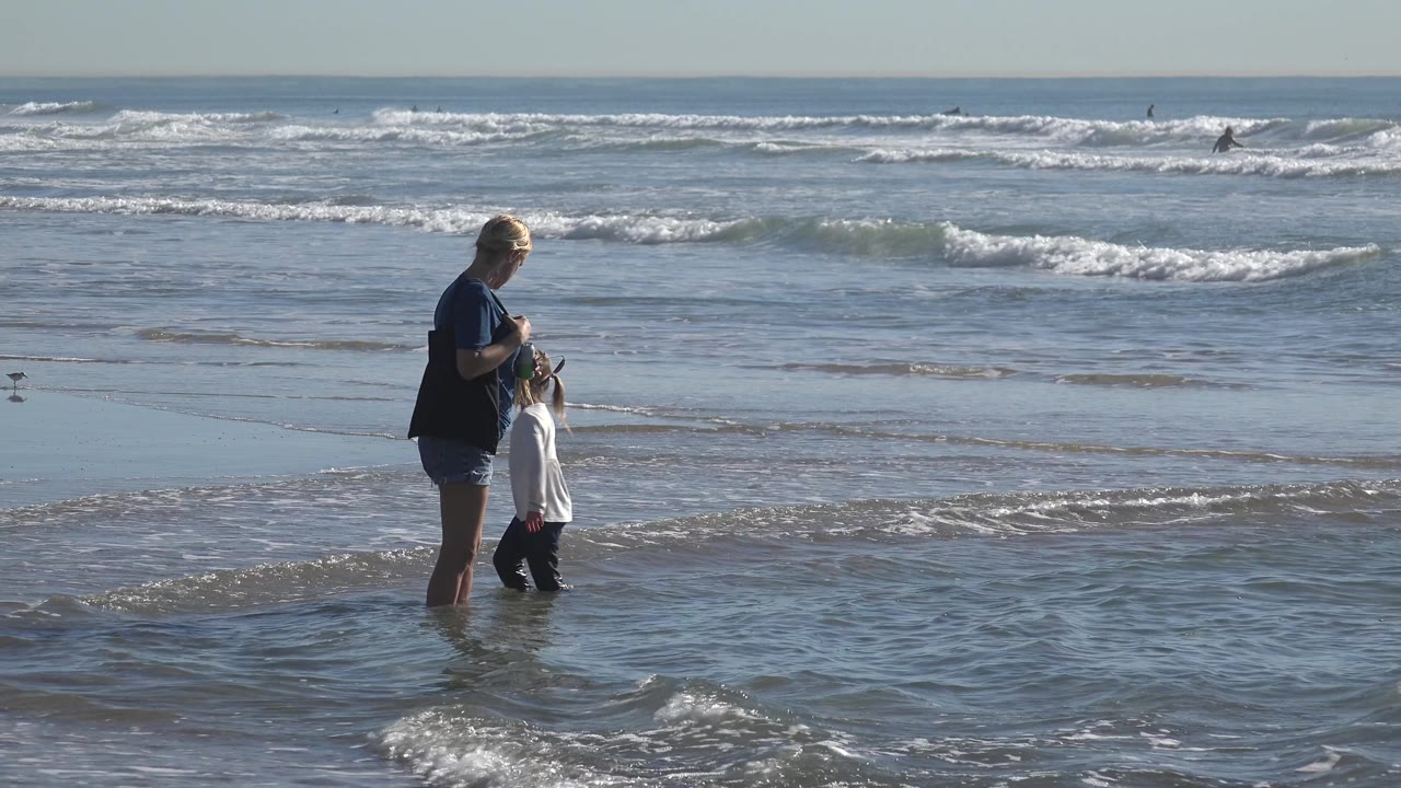At Huntington Beach, California, the grandmother bends slightly toward the child while small ripples and whitewater pass through the shallow foreground.