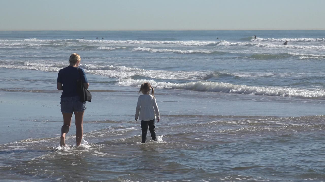 At Huntington Beach, California, an adult woman and young child stand in ankle-deep water as low waves roll toward the wet sand.