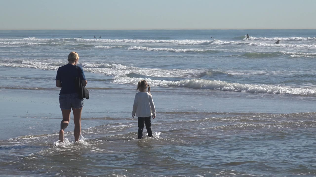 At Huntington Beach, California, the adult and child remain near the waterline while a small foamy wave spreads across the foreground.