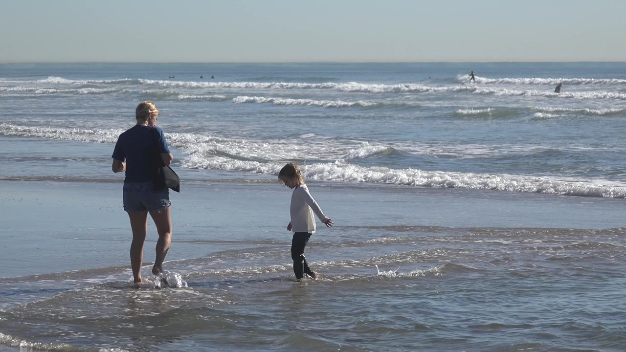 At Huntington Beach, California, the child moves to the right through shallow water as the adult stands a few steps away and watches the surf.