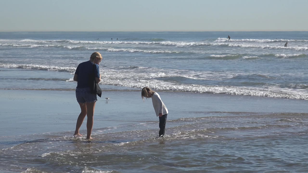 Child bends toward the water while an adult stands at the edge of the surf.