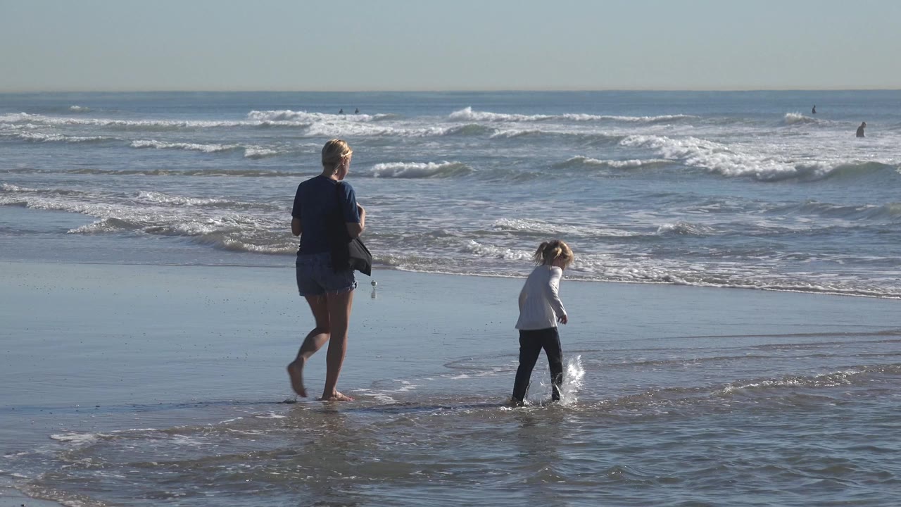 At Huntington Beach, California, an adult woman stands near the wet sand while a young child steps through shallow water with breaking waves behind them.
