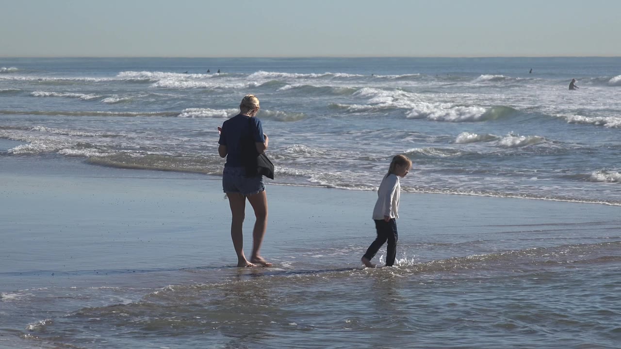 At Huntington Beach, the young child walks along the shallow surf to the right of the adult woman, with foamy waves and a clean ocean horizon filling the frame.
