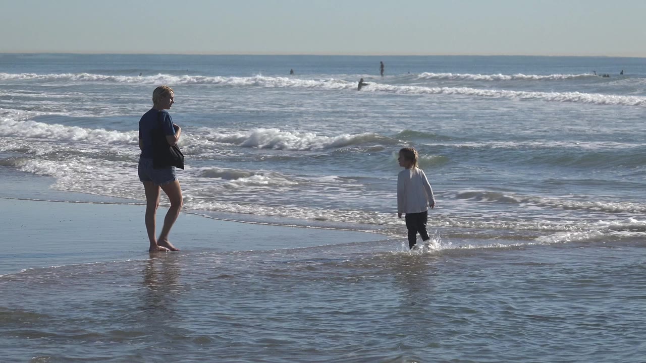 The woman stands barefoot in the wash while the child faces the surf at Huntington Beach, California, as a low wave breaks across the midground.