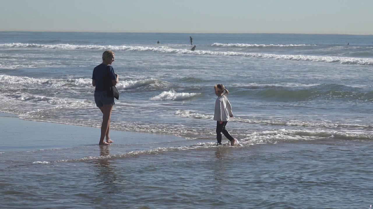 The child steps to the right through foamy water at Huntington Beach, California, while the woman remains near the shoreline with waves and distant surfers behind them.