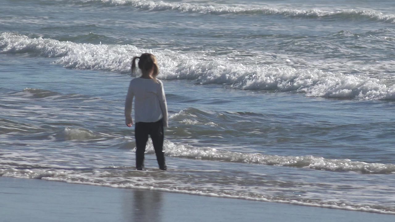 A young child stands near the edge of the water in Huntington Beach, California, facing small incoming waves with wet sand reflecting below.