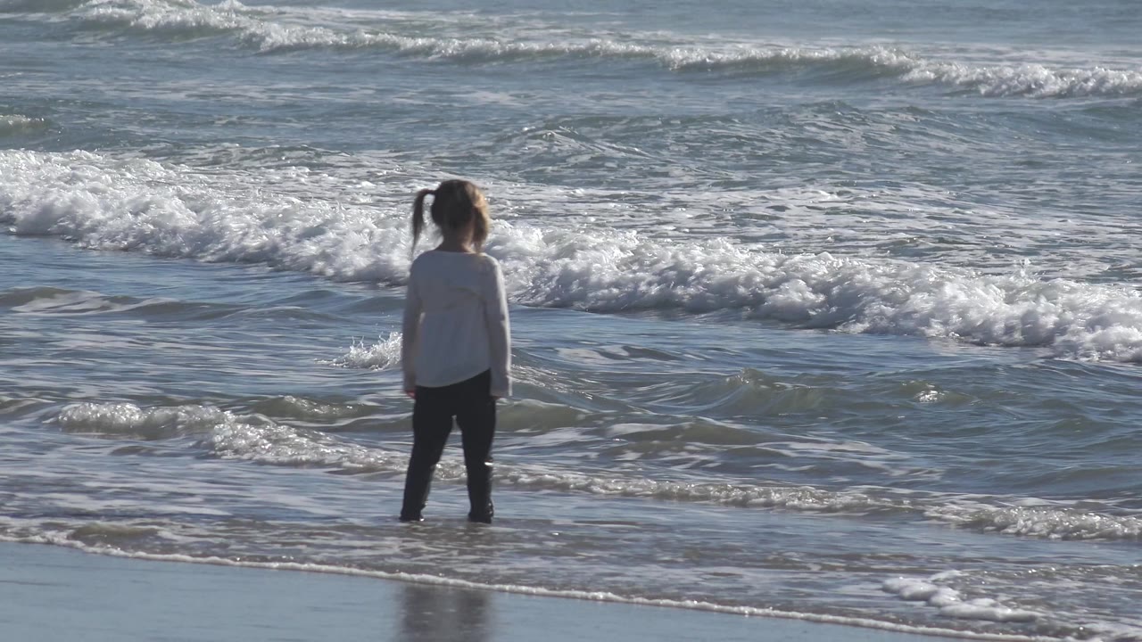 A side view shows the child near the Huntington Beach surf, turned slightly along the shore as foamy water moves across the background.