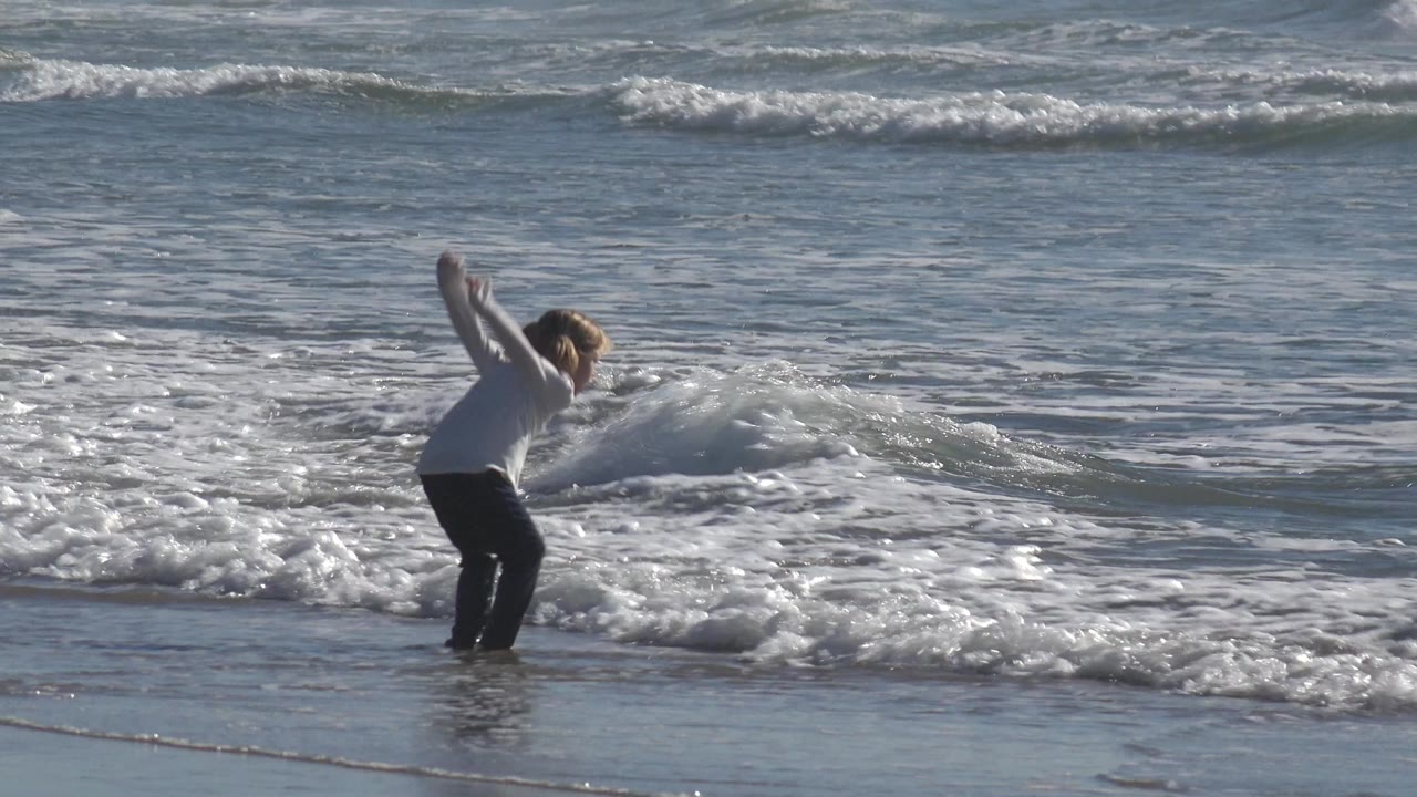 A young blonde girl in a light top and dark pants jumps near the edge of the surf at Huntington Beach, California, with foamy waves behind her.