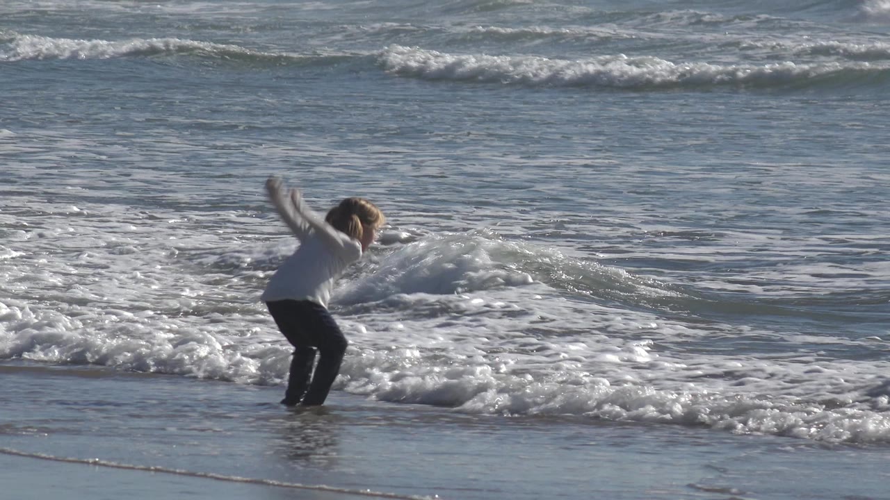 Young girl bends forward as surf reaches her feet.
