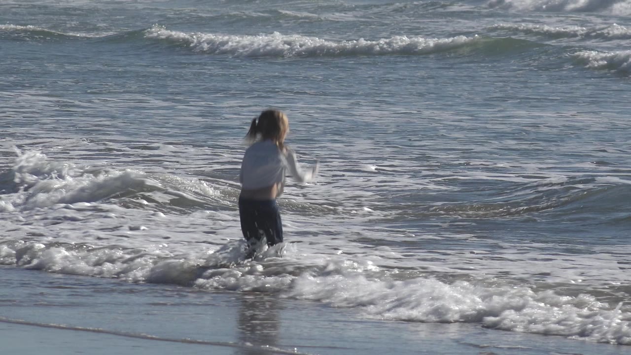 The child is turned toward the water at Huntington Beach as a small wave breaks in front of her and white foam spreads across the surface.