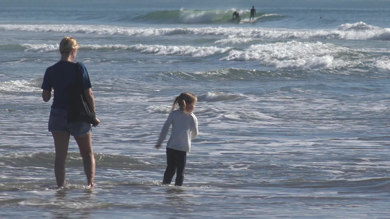 At Huntington Beach, California, an adult woman stands in ankle-deep water while a child in a white shirt faces the incoming surf, with surfers small in the background.