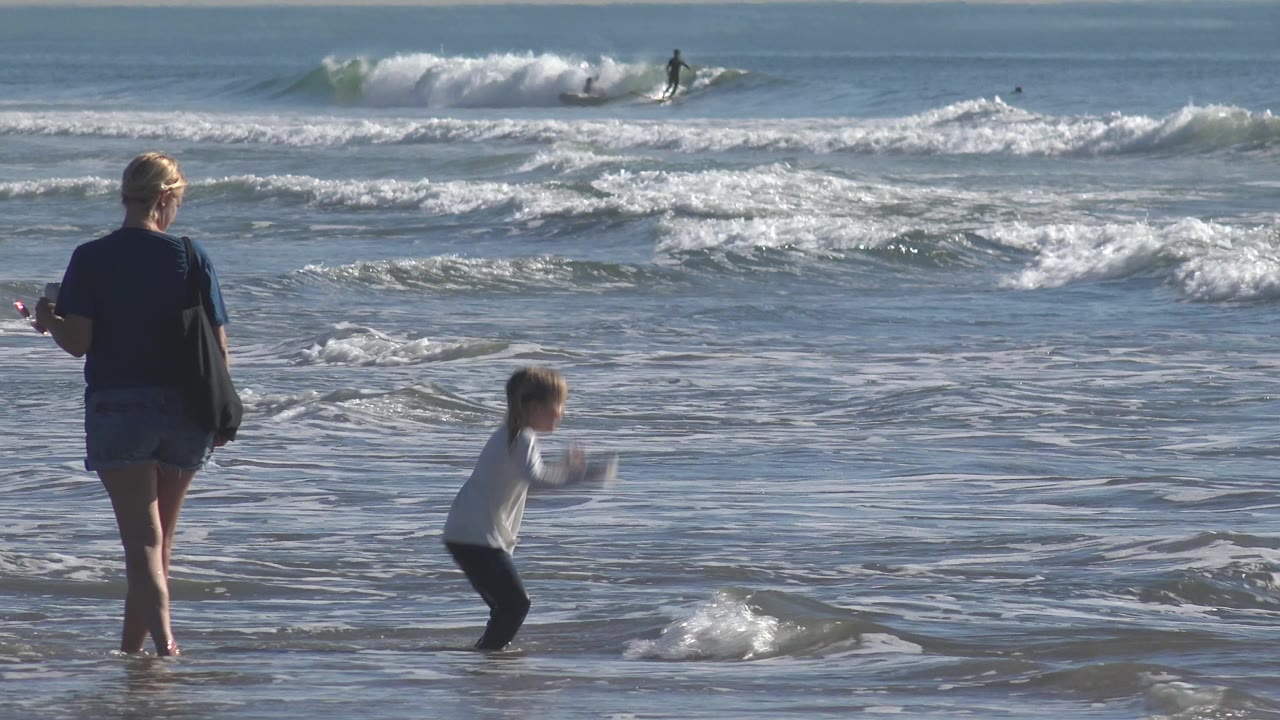 At Huntington Beach, California, the child lifts forward near a small breaking wave while the adult woman stands nearby, with brighter whitewater across the midground.
