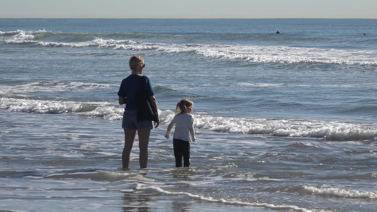 Adult and child near foamy water with surfers offshore.