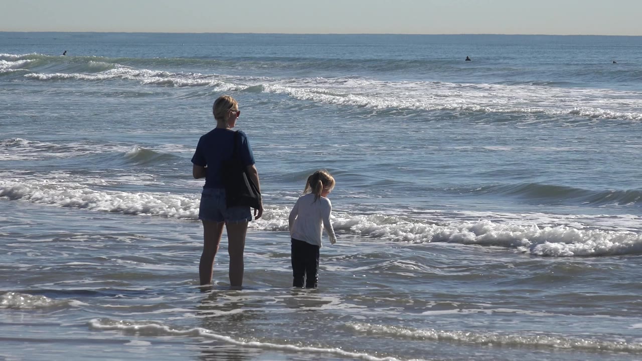 At Huntington Beach, California, the child shifts forward in the shallow water while the adult stands still, with low blue waves rolling in behind them.
