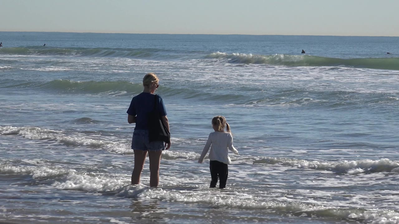 A woman and young child stand ankle-deep in the surf at Huntington Beach while small waves roll toward shore and distant surfers sit near the horizon.