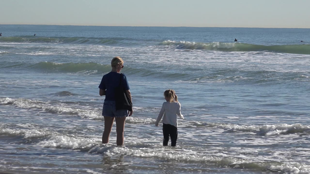 The woman and child remain in shallow Huntington Beach water, separated by a few feet as foam spreads around their legs and low waves rise behind them.