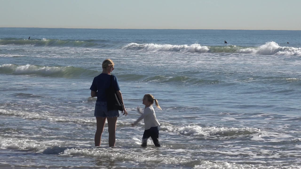 At Huntington Beach, the child turns toward the woman in the shallow water while a white-capped wave breaks in the background with small surfer silhouettes offshore.