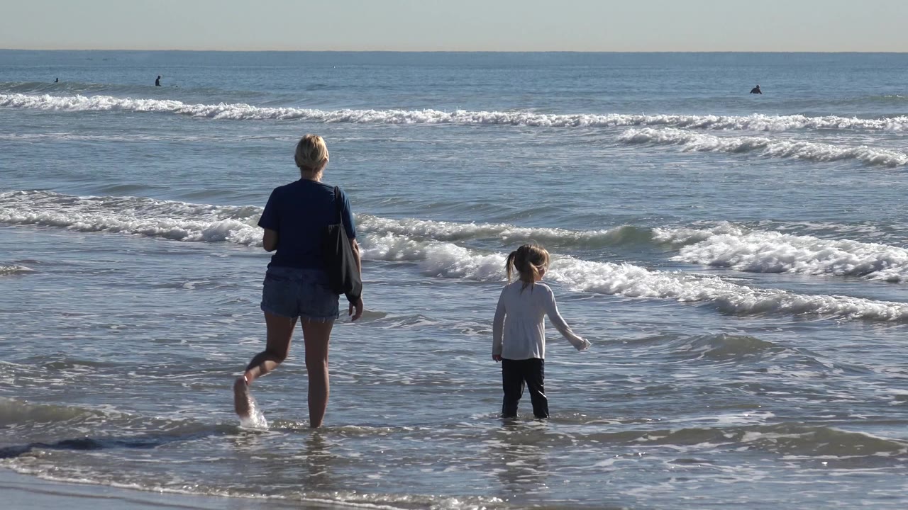 At Huntington Beach, California, a woman and young child walk through ankle-deep water while white surf rolls across the background.