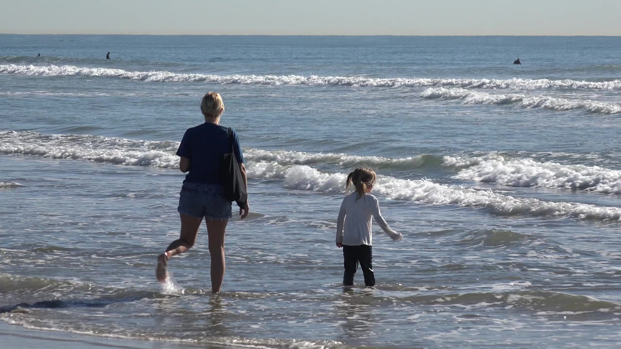 Woman stands in surf while child steps ahead near small waves.