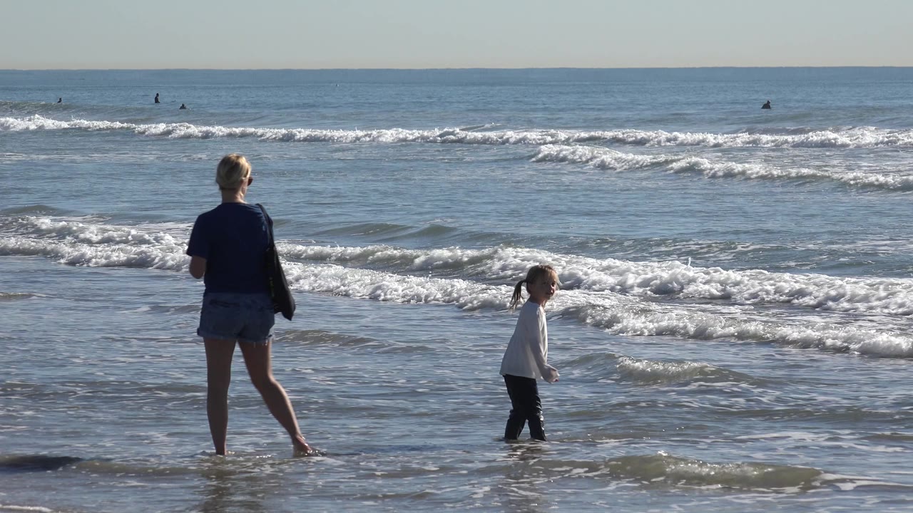 At Huntington Beach, California, the child stands closer to the foamy water as the woman trails behind with a shoulder bag and the surf line stretches across the frame.