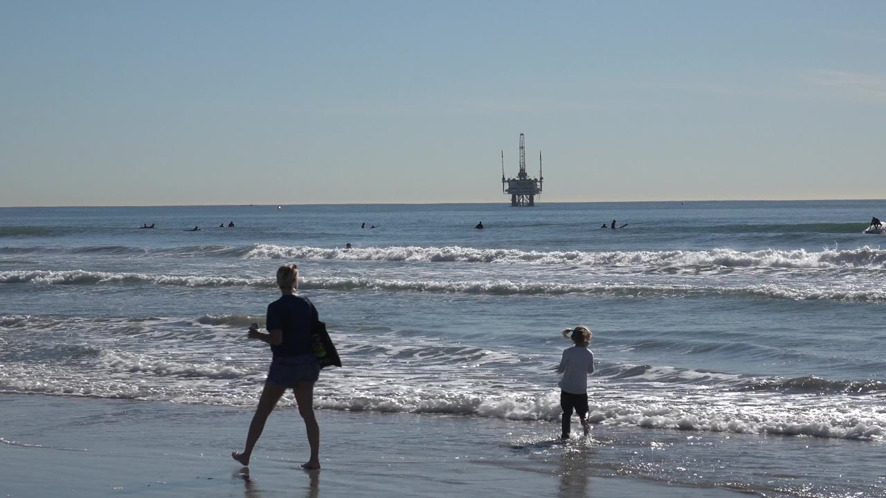 At Huntington Beach, an adult stands on wet sand while a child faces the incoming shore break, with surfers and an offshore platform visible beyond the waves.