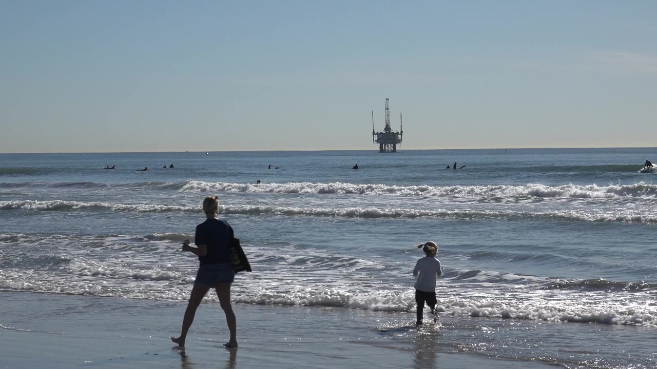 At Huntington Beach, the child remains in the shallow water as an adult walks along the reflective beach, with whitewater and surfers spread across the background.