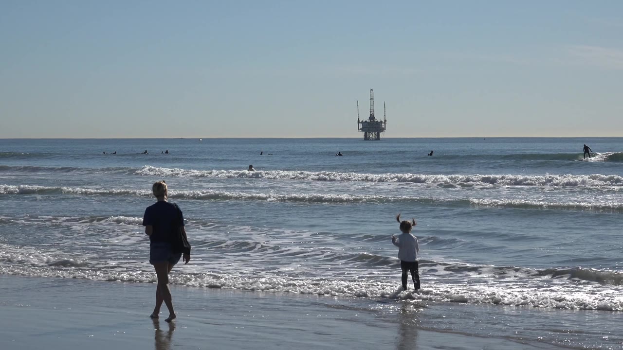 At Huntington Beach, the adult moves across the foreground while the child stands near foamy water, with small figures of surfers and an offshore platform on the horizon.