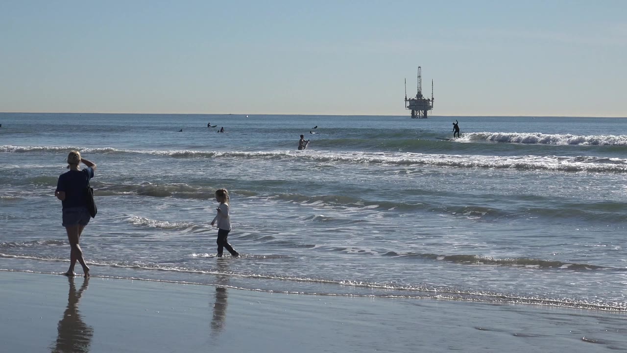 A parent and child walk near the shoreline at Huntington Beach, California, with small waves, surfers, and an offshore platform on the horizon.