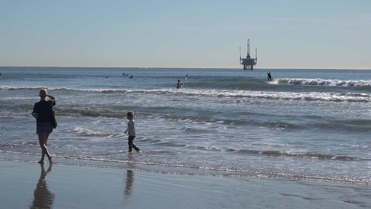 At Huntington Beach, California, the parent stands near the reflective wet sand as the child walks through shallow foam with surfers beyond the break.