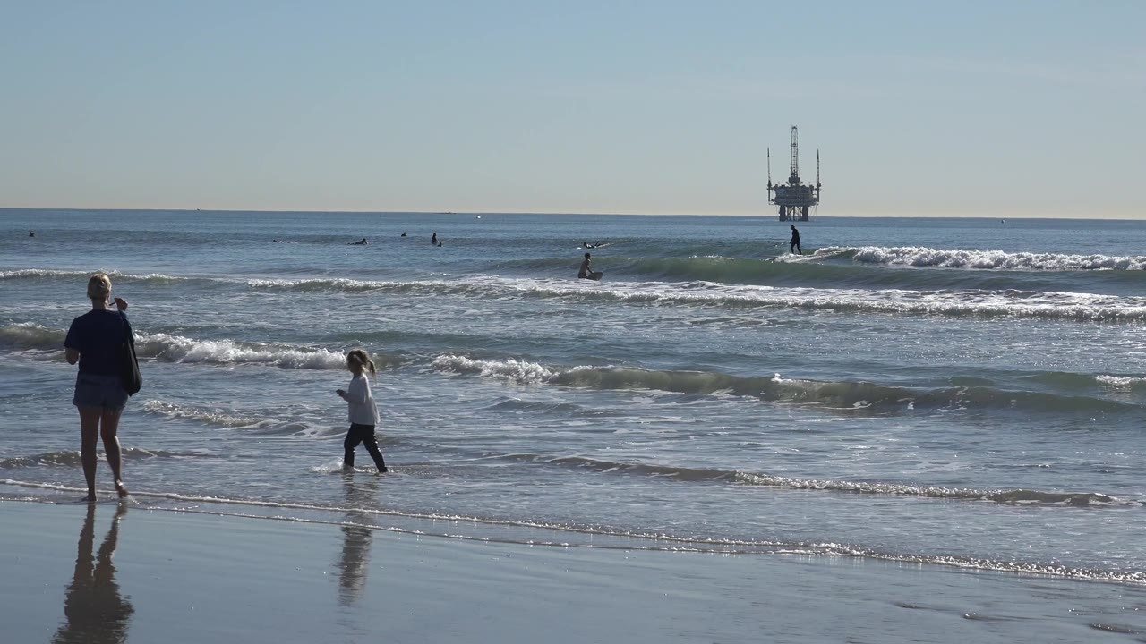 The parent and child remain near the Huntington Beach waterline as a low wave breaks behind them and surfers spread across the midground.