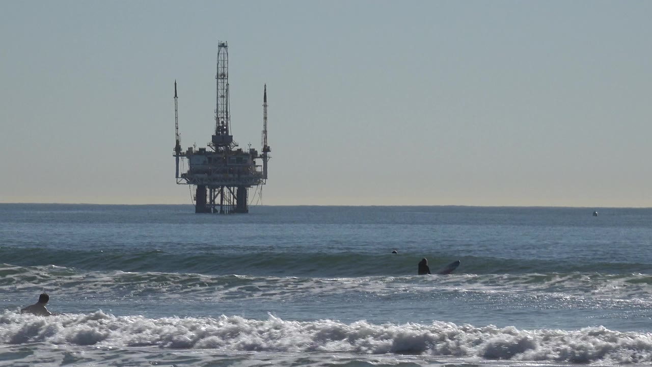 Huntington Beach surfers are visible in low surf while a dark offshore oil platform stands near the horizon under a pale sky.