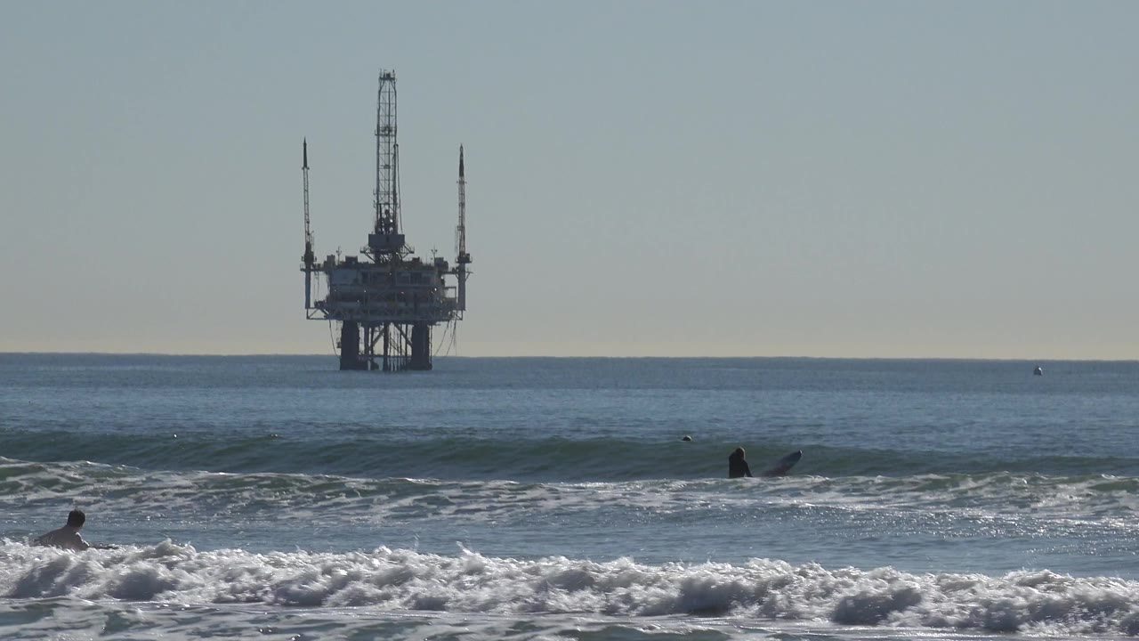 A surfer moves through foreground whitewater at Huntington Beach as the offshore oil platform remains centered in the distance.