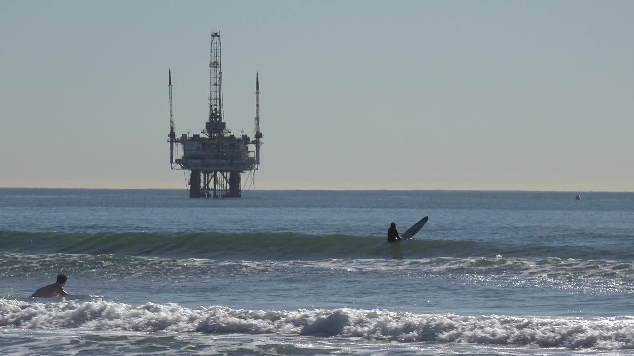 Multiple surfers are spaced across the Huntington Beach waterline as a small swell rises in front of the distant oil platform.