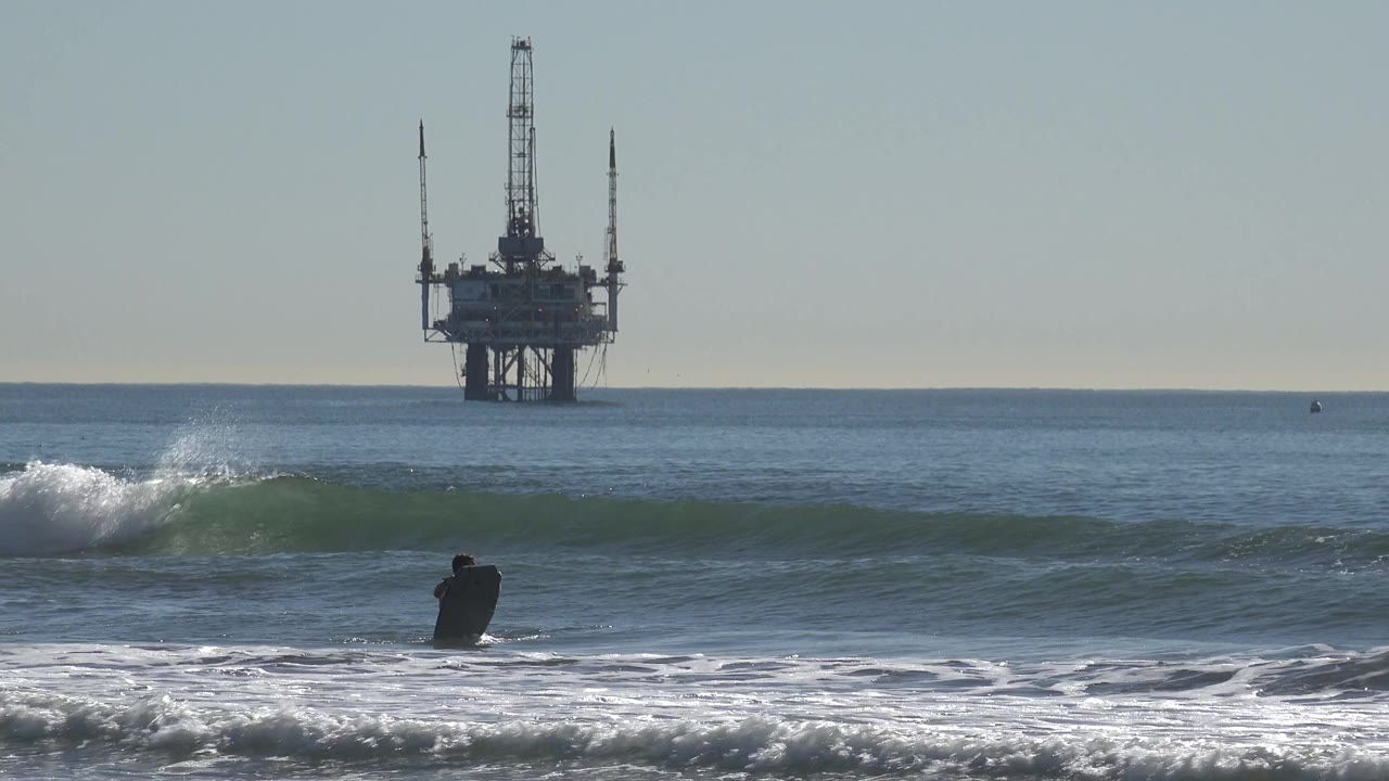 At Huntington Beach, California, the surfer sits low in the foreground water as a green wave lifts across the frame and the oil rig remains centered beyond the horizon.