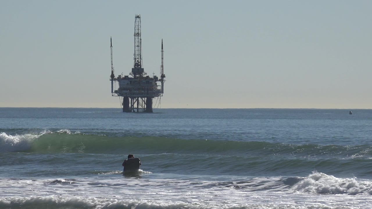 At Huntington Beach, California, the surfer is farther right in the shore break as the wave crest starts to spill and the offshore oil rig sits on the background horizon.