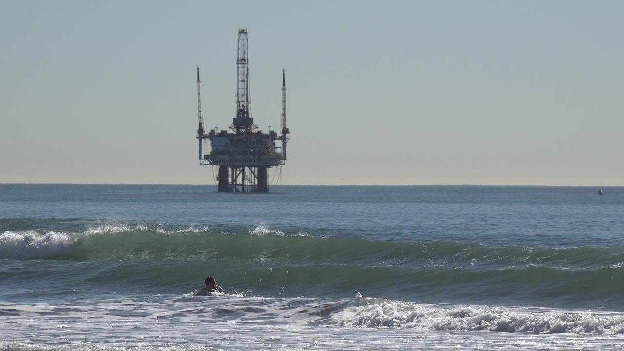 Oil rig beyond calm water as a surfer appears low in the lower left surf.