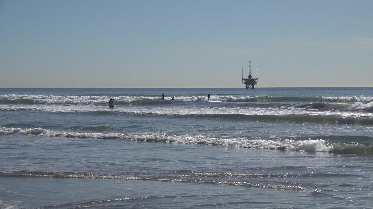 Huntington Beach surf rolls toward wet sand while distant people stand or move in the water and a dark offshore platform sits near the horizon.