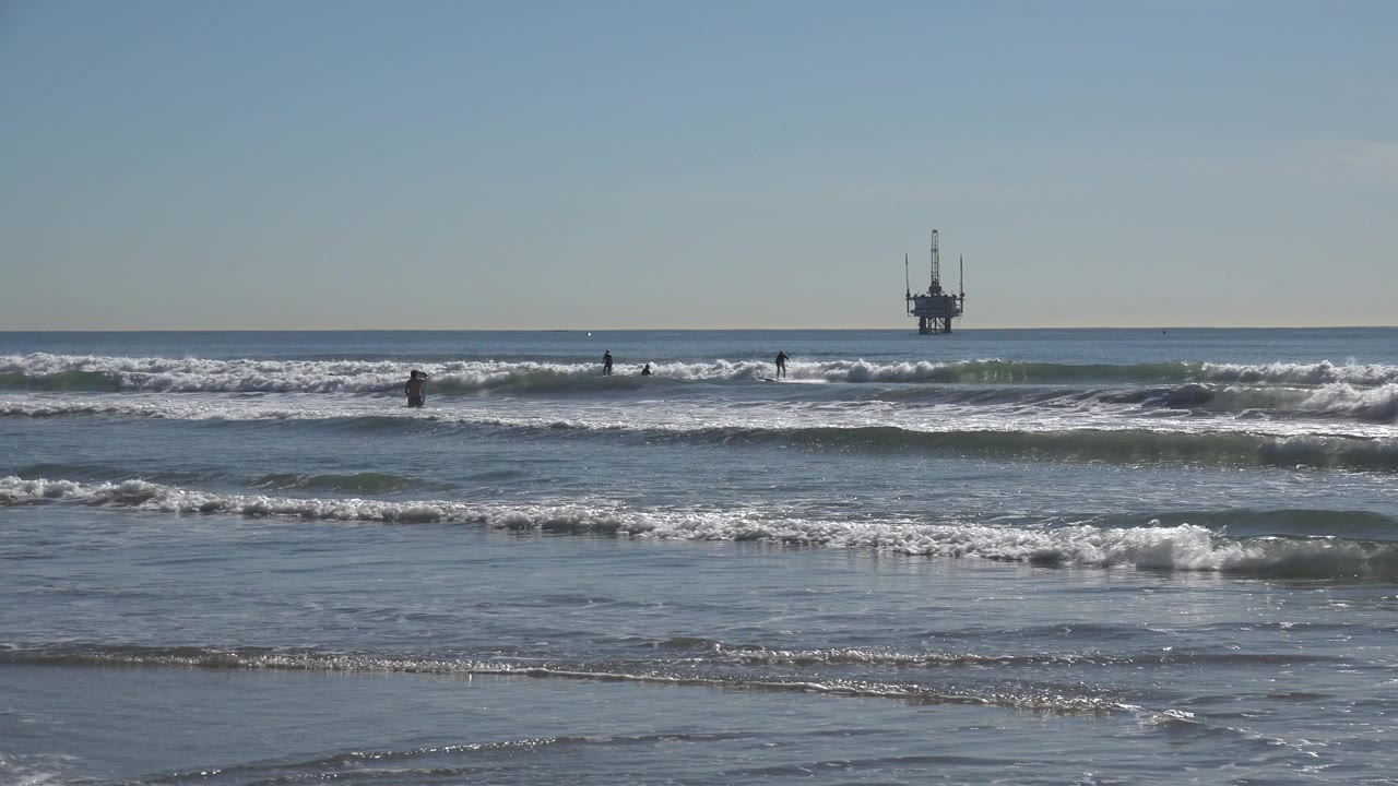 At 0:01, Huntington Beach waves form bright white bands as several distant people remain scattered across the midground water.