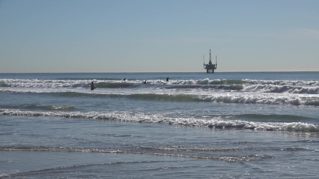 At 0:02, a larger breaking wave crosses the Huntington Beach midground while distant people appear as dark silhouettes against the water.