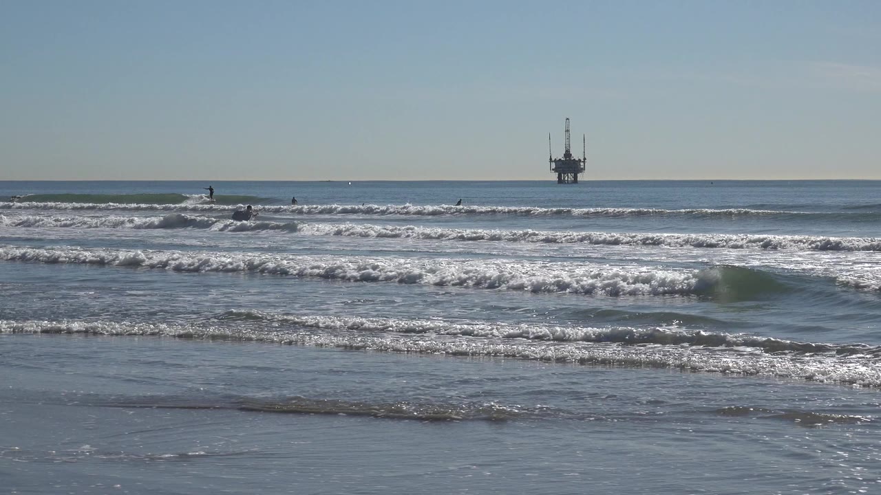Huntington Beach surf breaks across wet sand while a distant offshore oil rig sits near the center of the horizon.