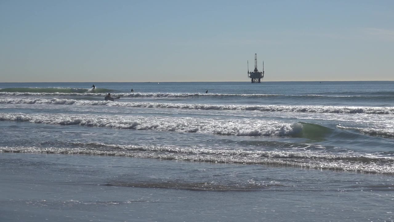 At Huntington Beach, small distant people appear beyond the shore break while a dark oil platform stands on the right side of the horizon.
