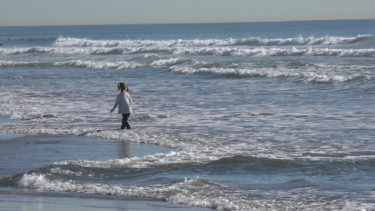 At Huntington Beach, California, a young girl in a light top walks left across shallow water while white surf breaks behind her.