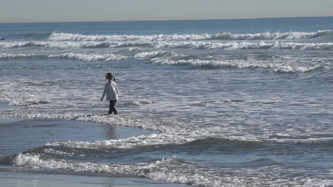 At Huntington Beach, California, the girl has moved toward the center of the frame, standing in reflective shallows with rows of white waves behind her.